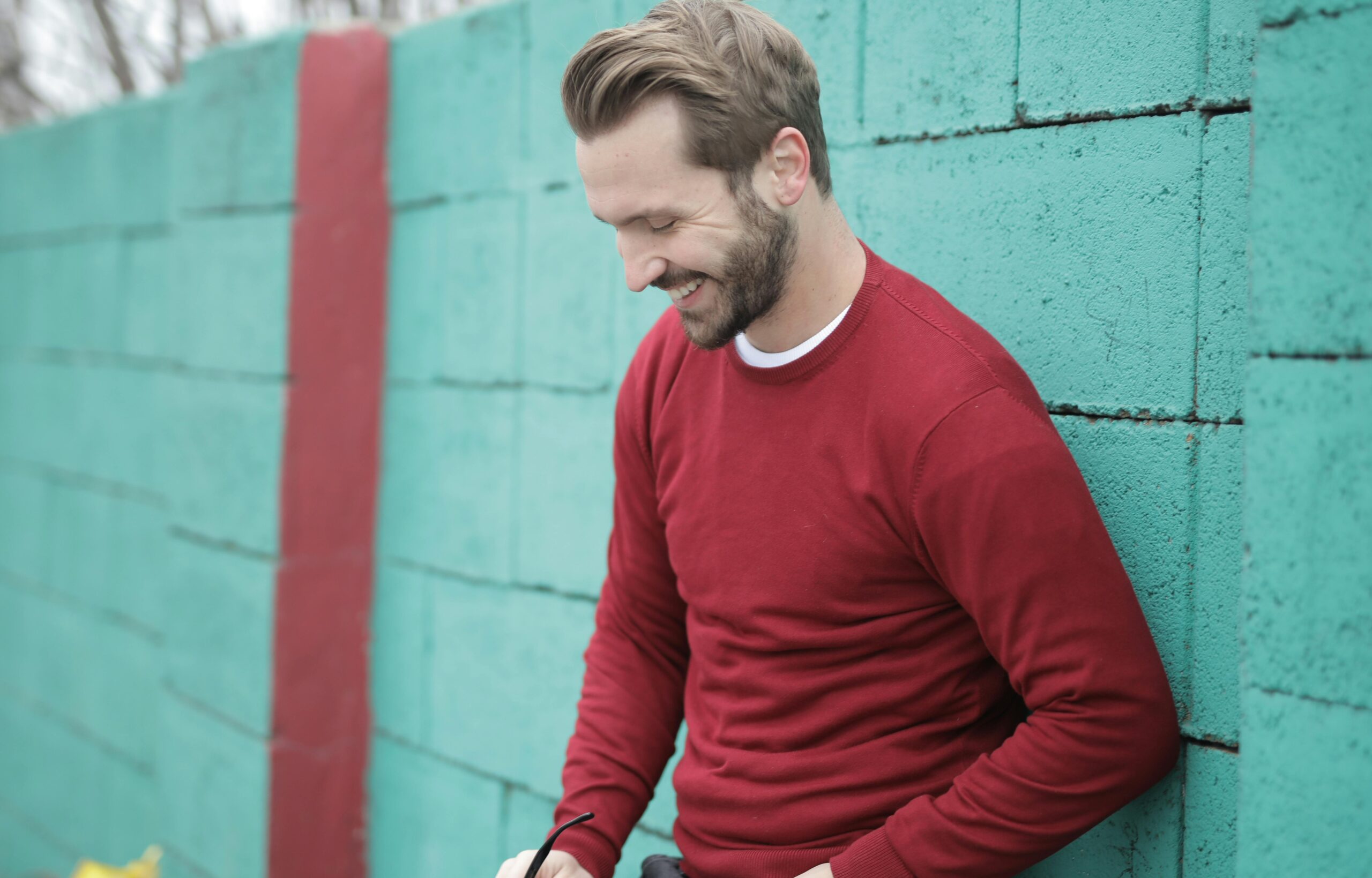 Home A man in a red sweater smiles while leaning against a colorful green wall outdoors.
